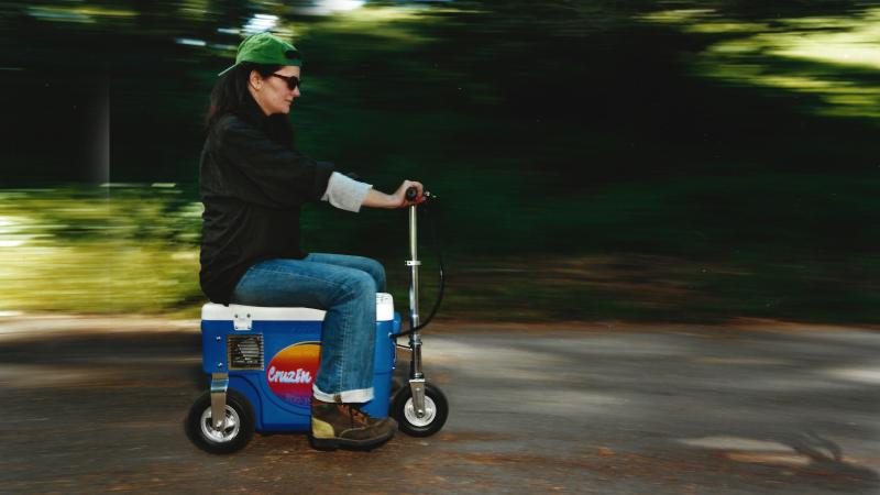 A woman is riding on a vehicle that looks like a cooler with wheels from the film Do I Need This?